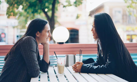 Two women talk across an outdoor cafe table.