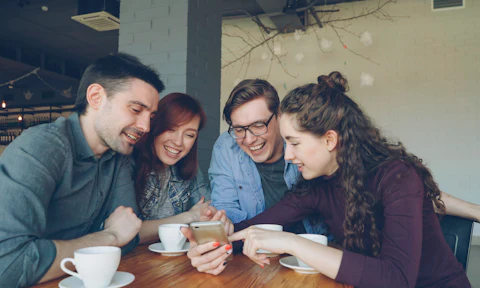 Four friends at a cafe table look at one phone together and laugh.