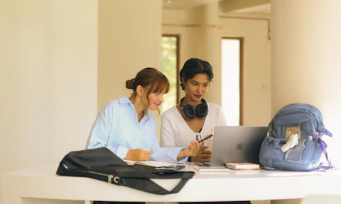 Two women read together at a table with a laptop.