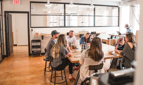 Six people talk around a long wooden table in a bright room.